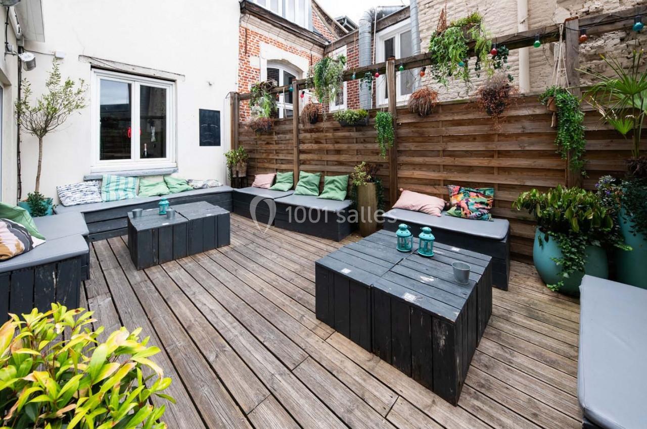 Terrasse en bois avec bancs et tables noires, coussins colorés, plantes suspendues et pots verts.
