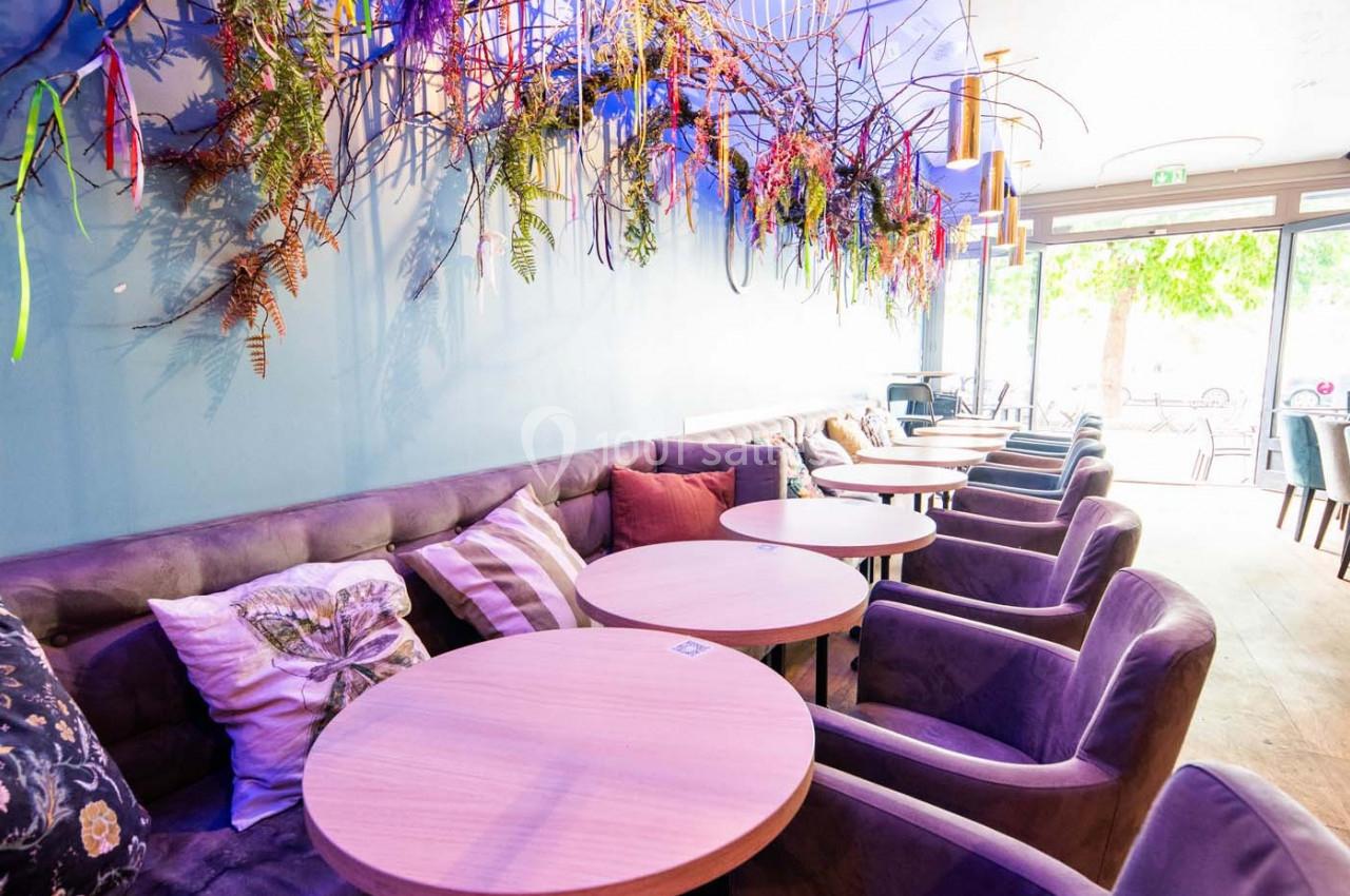Banquettes violettes et tables rondes en bois dans un café décoré de plantes suspendues et éclairé par une grande baie…