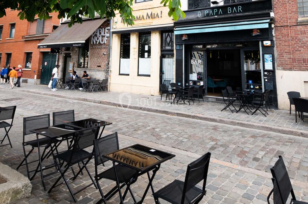 Terrasse vide avec tables et chaises noires sur une rue pavée, devant des façades de cafés et restaurants.