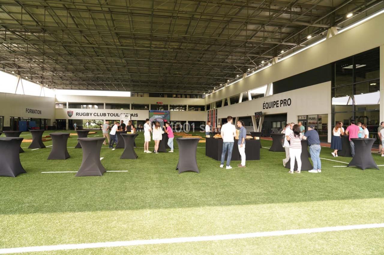 Personnes rassemblées dans un espace intérieur sur gazon synthétique, avec des tables hautes noires disposées autour.