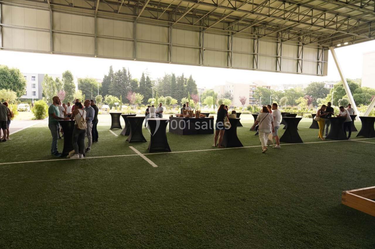 Des personnes discutent autour de tables hautes sous un espace couvert, avec un terrain en gazon synthétique.