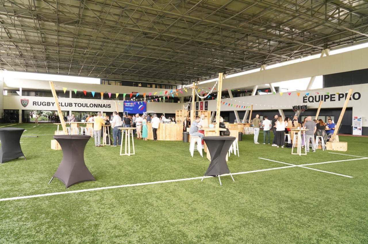 Personnes rassemblées dans un espace couvert sur gazon, avec des tables hautes et des décorations festives.