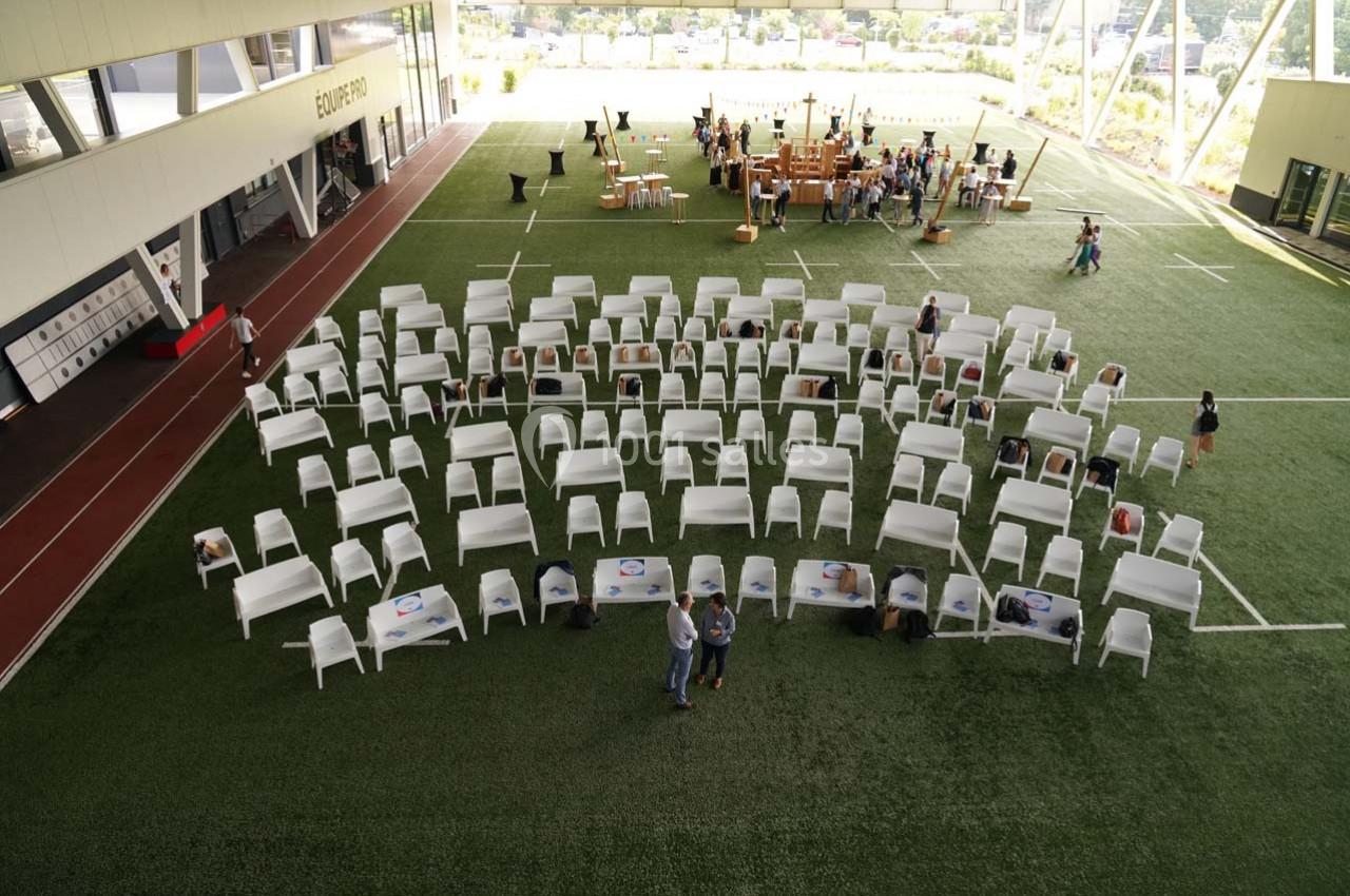 Chaises blanches disposées en rangées sur un terrain synthétique, avec des tables et des personnes en arrière-plan.