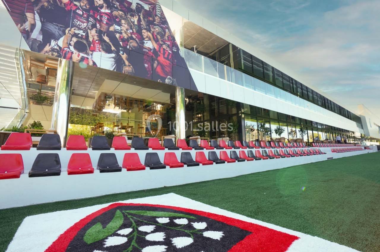 Tribune extérieure avec des sièges rouges et noirs devant un bâtiment moderne, sur une pelouse ornée d'un motif floral.