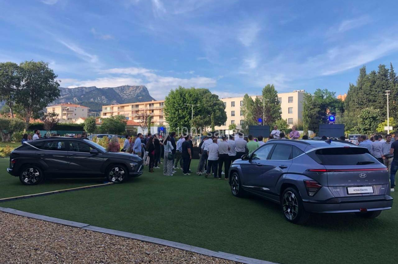 Deux SUV gris exposés sur une pelouse, avec un groupe de personnes rassemblées en arrière-plan dans un cadre urbain.