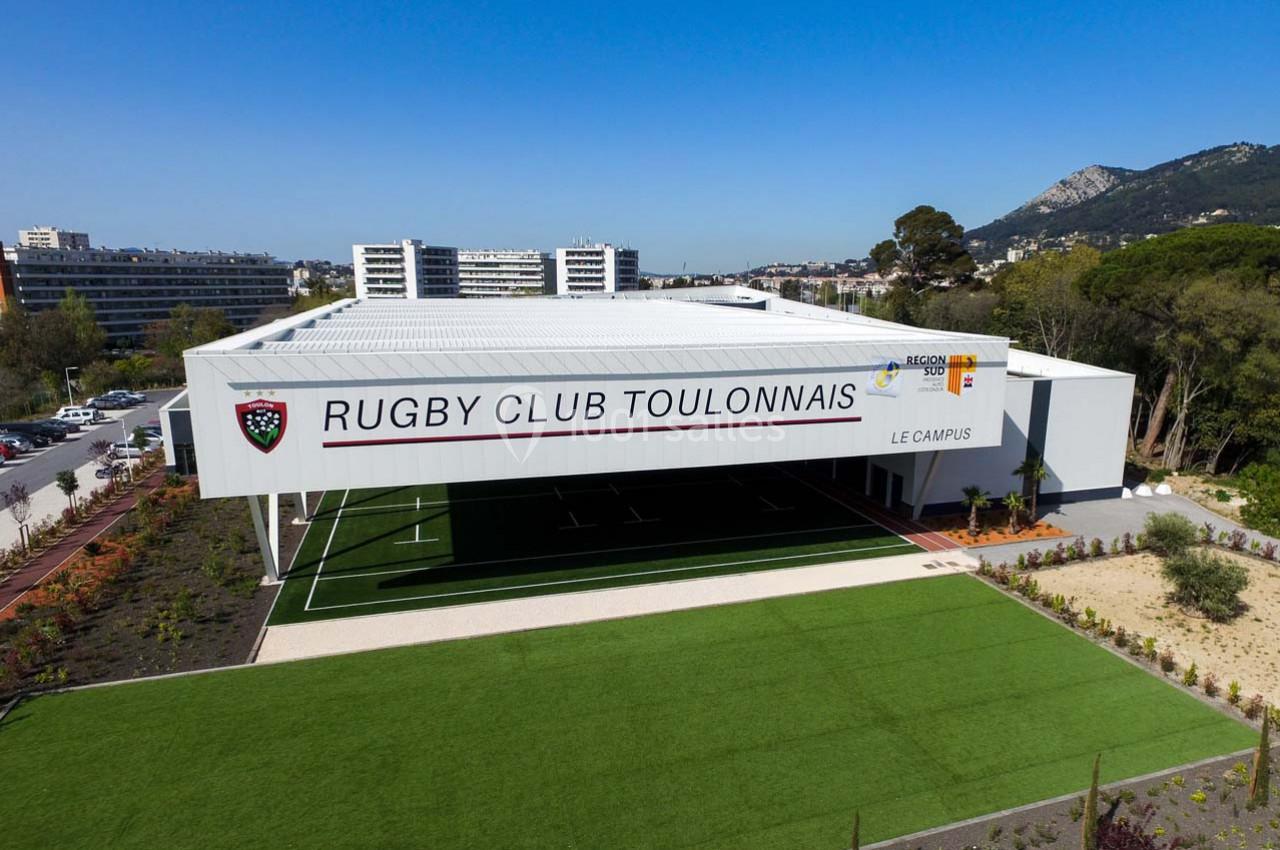 Vue extérieure du bâtiment du Rugby Club Toulonnais avec terrain synthétique et paysage urbain en arrière-plan.