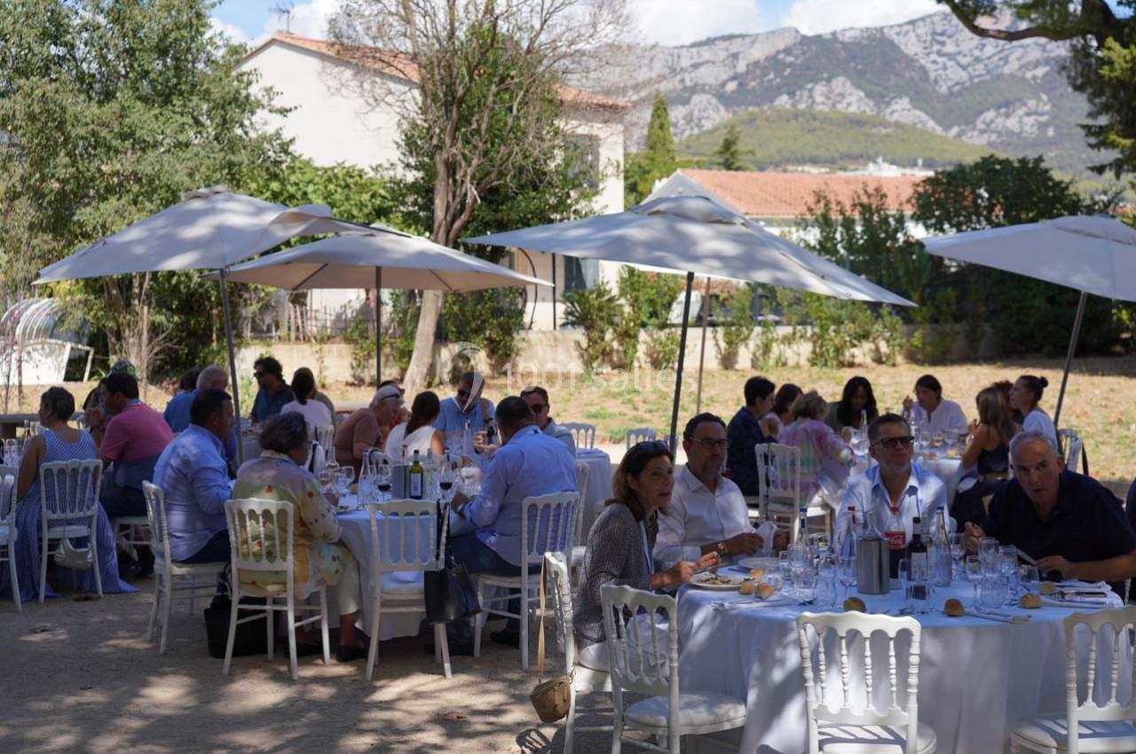 Des convives assis à des tables rondes en plein air, sous des parasols, dans un jardin avec des montagnes en arrière-plan.