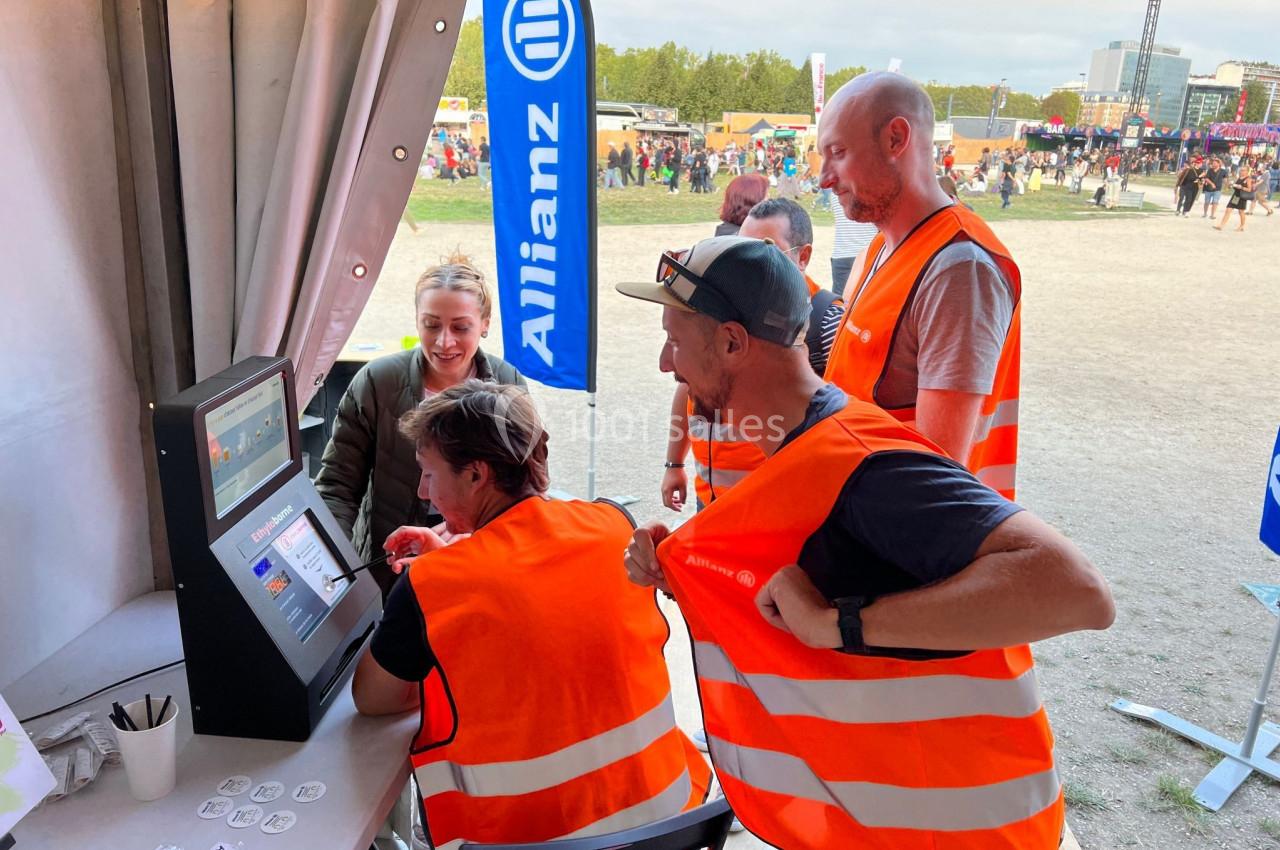 Des bénévoles en gilets orange utilisent une borne interactive sous une tente lors d'un événement en plein air.