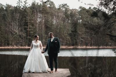 Un couple en tenue de mariage se tient sur un ponton en bois devant un lac entouré d'arbres.