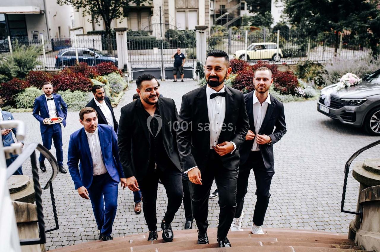 Un groupe d'hommes en costume monte des marches dans un cadre urbain avec des voitures et de la végétation en arrière-plan.