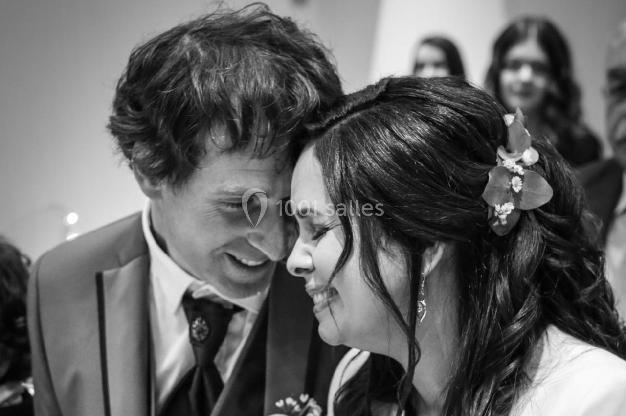 Un couple souriant, front contre front, lors d'une cérémonie, entouré de quelques personnes en arrière-plan flou.