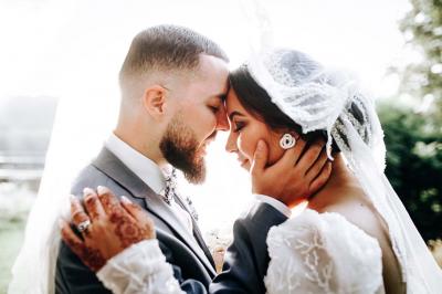 Un couple en tenue de mariage pose devant une voiture ancienne noire et un bâtiment historique entouré de verdure.