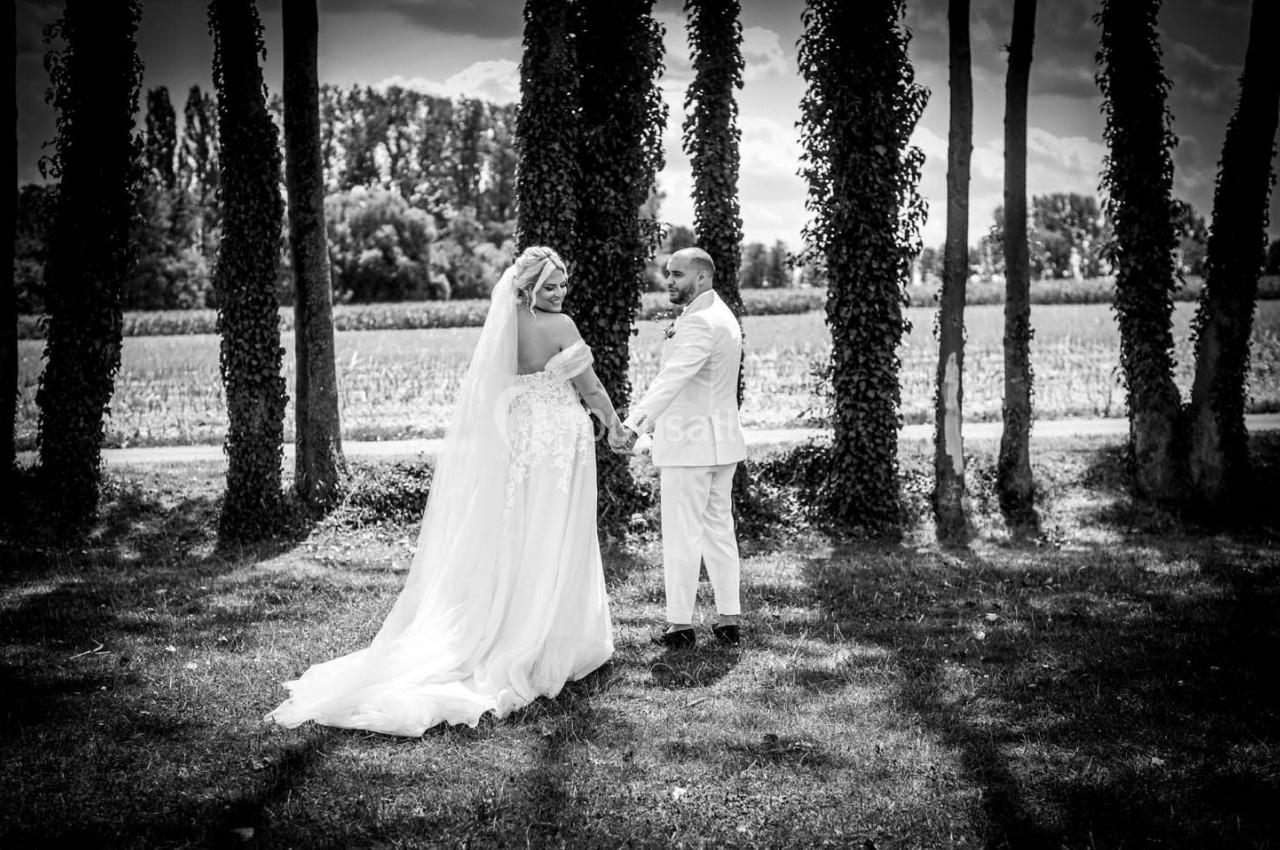Un couple de mariés se tient la main dans un parc ombragé, entouré d'arbres, sous un ciel légèrement nuageux.