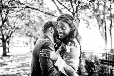 Un couple en tenue de mariage pose devant une voiture ancienne noire et un bâtiment historique entouré de verdure.