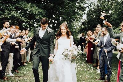 Un couple en tenue de mariage pose devant une voiture ancienne noire et un bâtiment historique entouré de verdure.