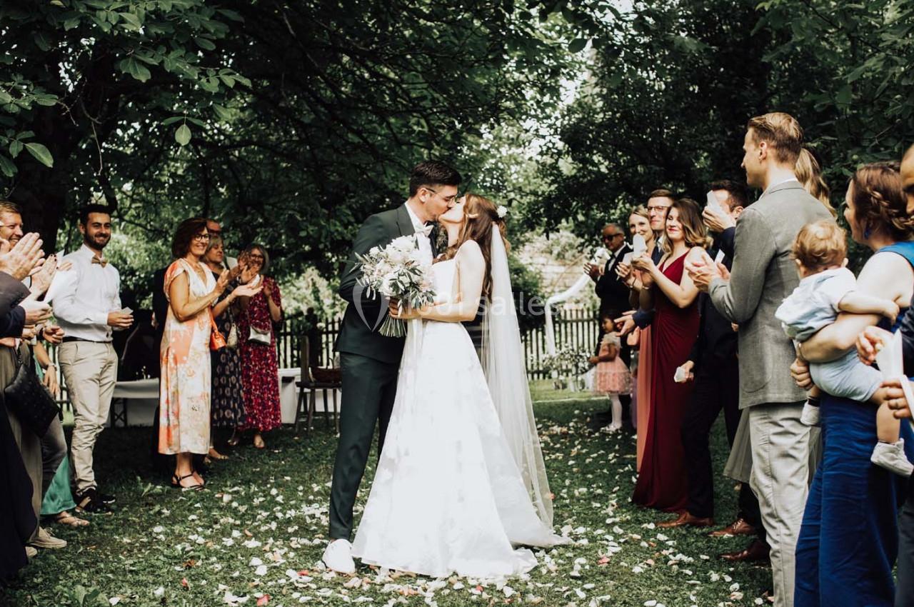Un couple de mariés s'embrasse dans un jardin entouré d'invités applaudissant sous les arbres.