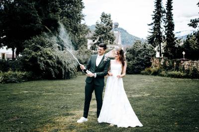 Un couple en tenue de mariage pose devant une voiture ancienne noire et un bâtiment historique entouré de verdure.