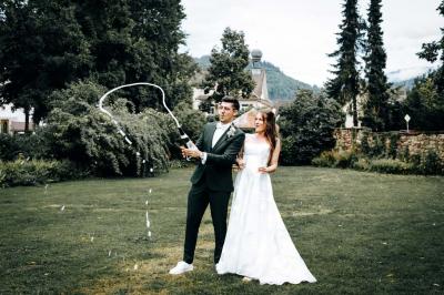 Un couple en tenue de mariage pose devant une voiture ancienne noire et un bâtiment historique entouré de verdure.
