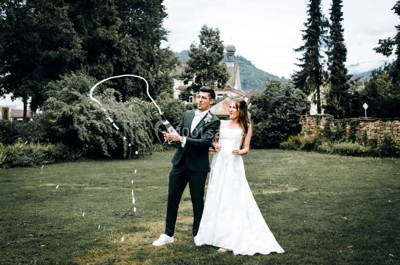 Un couple en tenue de mariage dans un jardin, l'homme ouvrant une bouteille de champagne qui éclabousse.