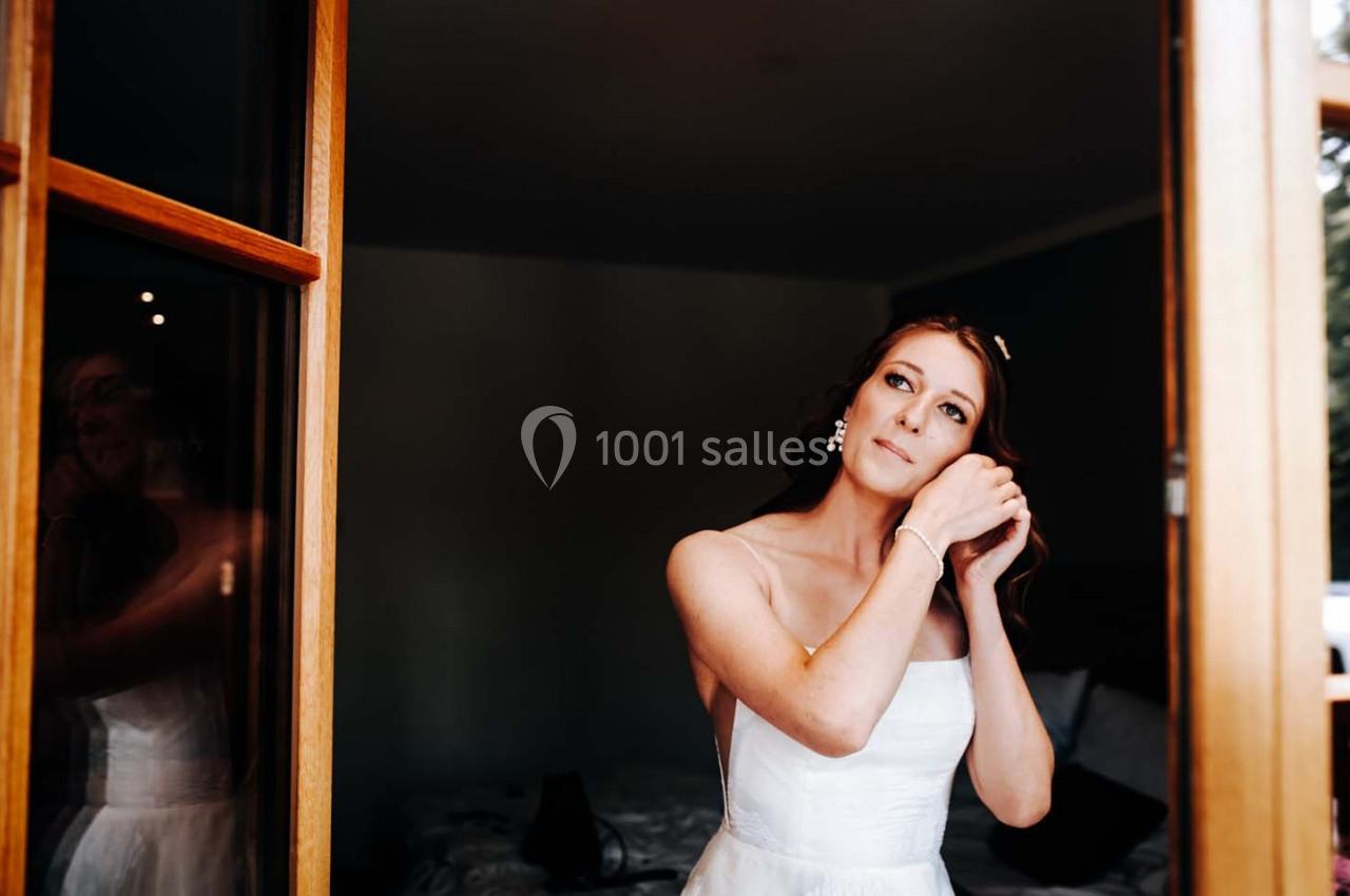 Une femme en robe blanche ajuste ses boucles d'oreilles devant une fenêtre ouverte dans une pièce sombre.