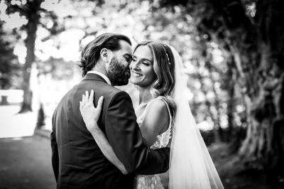 Un couple en tenue de mariage pose devant une voiture ancienne noire et un bâtiment historique entouré de verdure.