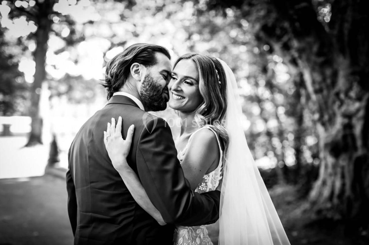 Un couple en tenue de mariage s'enlace tendrement dans un parc, capturé en noir et blanc.