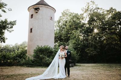 Un couple en tenue de mariage pose devant une voiture ancienne noire et un bâtiment historique entouré de verdure.