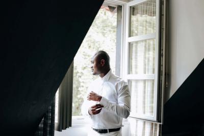 Un couple en tenue de mariage pose devant une voiture ancienne noire et un bâtiment historique entouré de verdure.