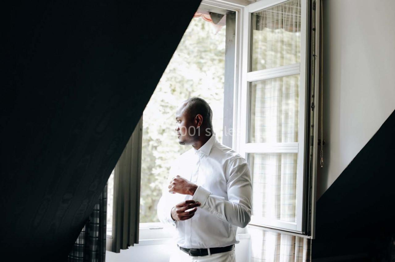 Homme en chemise blanche regardant par une fenêtre ouverte, baigné par une lumière naturelle.