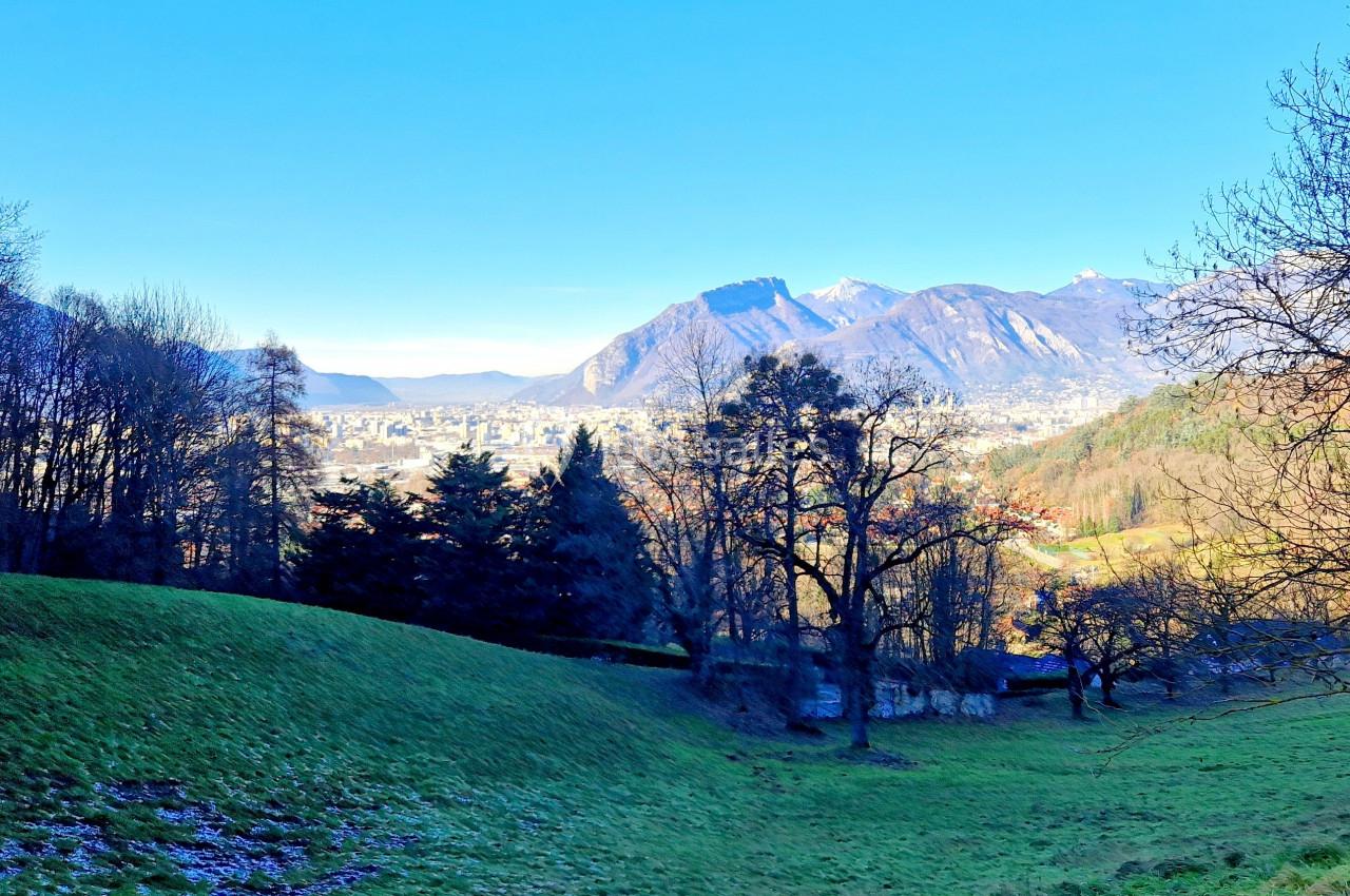 Vue d'une vallée verdoyante avec des arbres, des montagnes en arrière-plan et une ville visible au loin sous un ciel bleu.