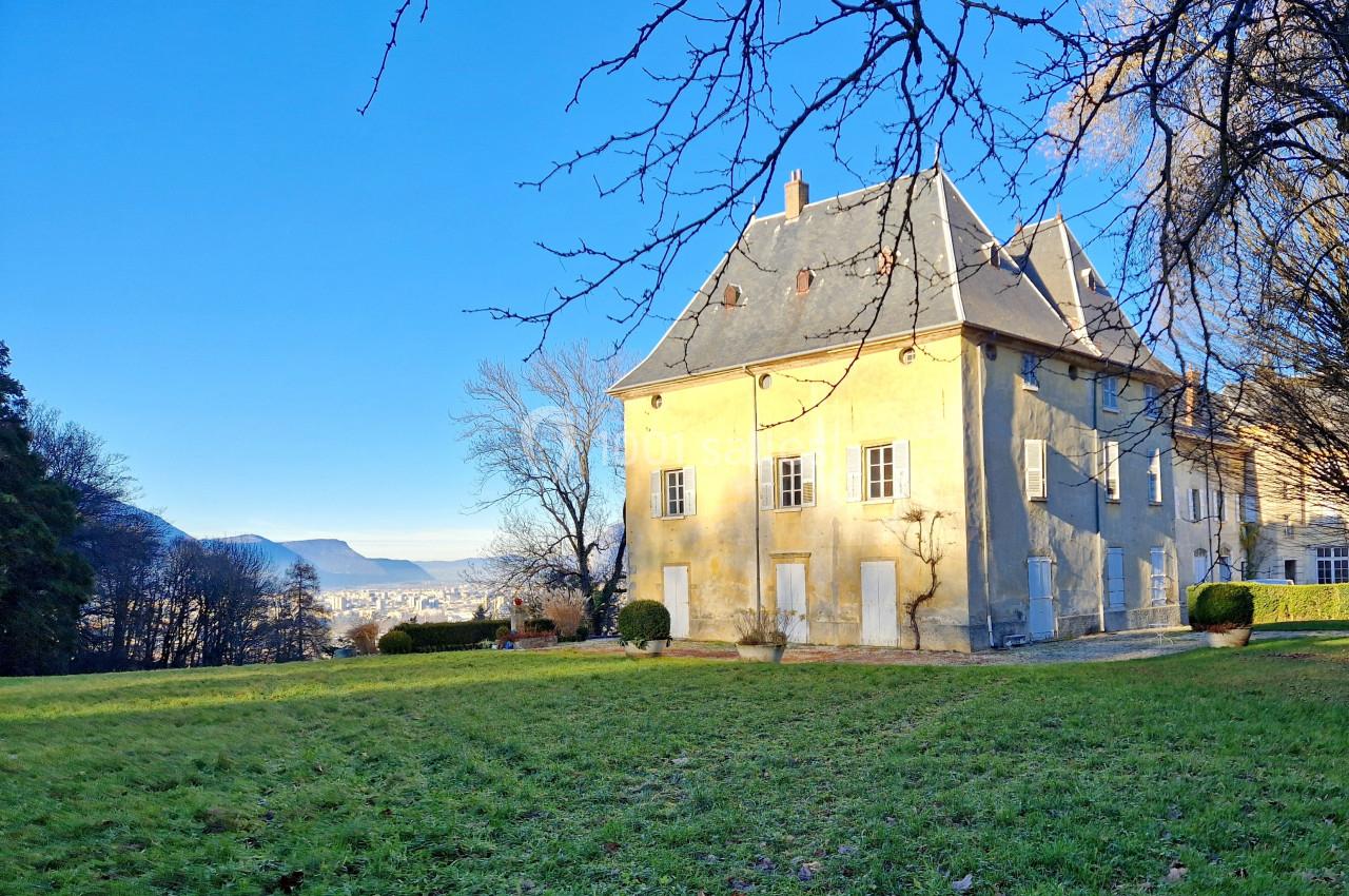 Grande maison ancienne avec toit en ardoise, entourée d'un jardin et d'arbres, sous un ciel bleu clair.