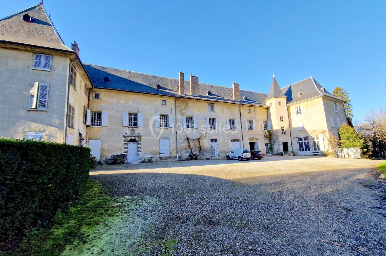 Façade d'un château ancien avec toit en ardoise, cour gravillonnée et végétation en bordure, sous un ciel dégagé.