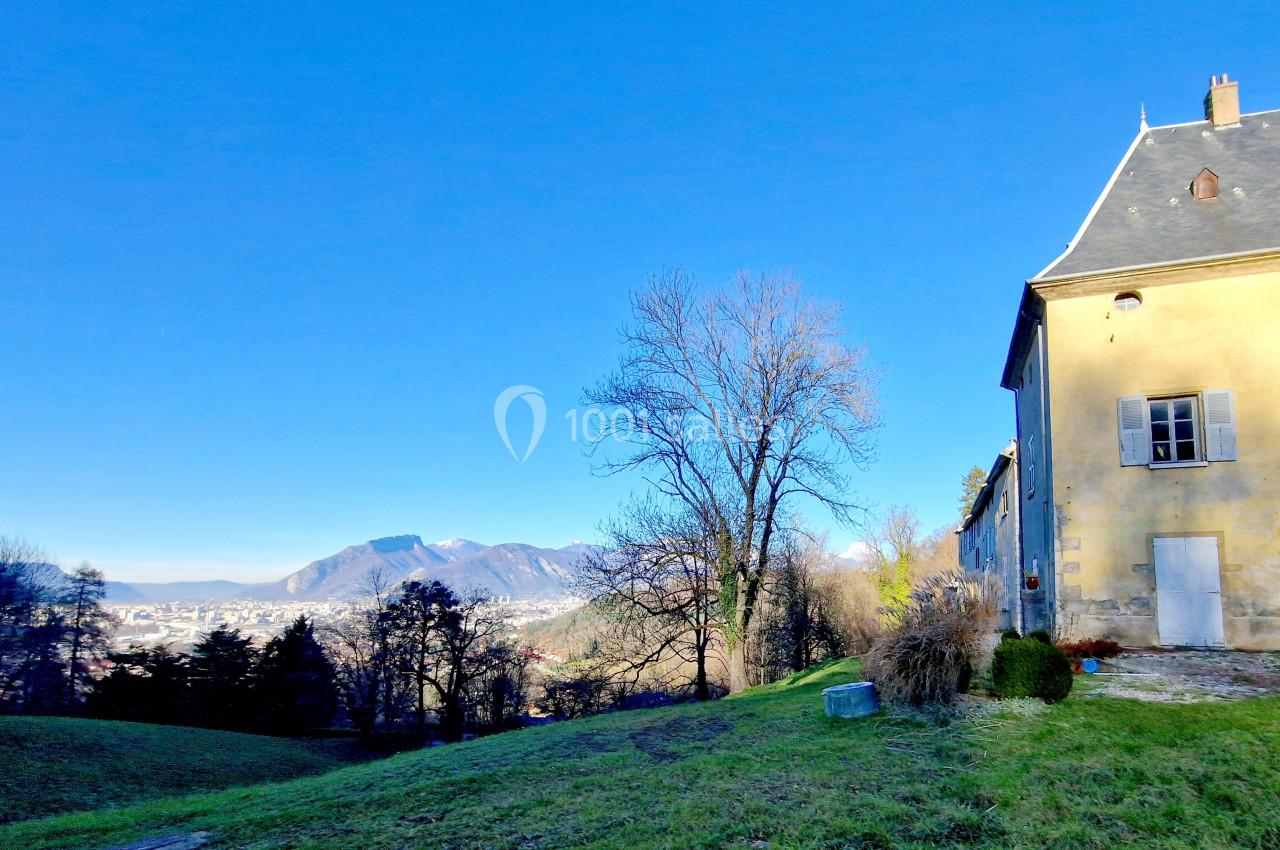 Vue d'un bâtiment ancien en pierre près d'une pelouse, avec des montagnes et un ciel bleu en arrière-plan.