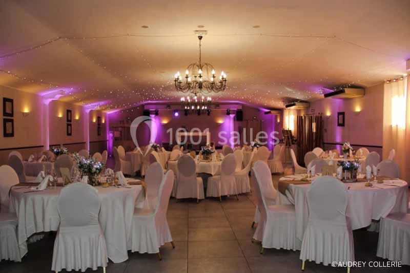Salle de réception décorée avec des tables rondes, nappes blanches, éclairage tamisé et guirlandes lumineuses au plafond.