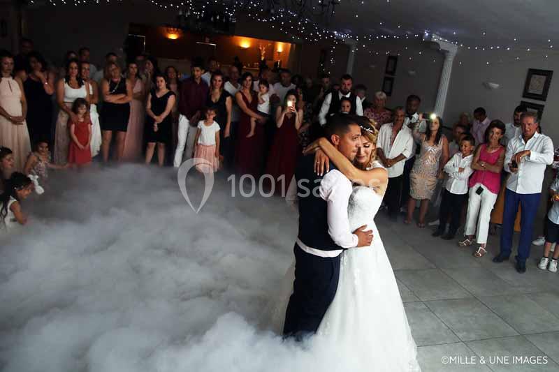 Un couple de mariés danse au centre d'une salle entourée d'invités, avec un sol recouvert de fumée légère.