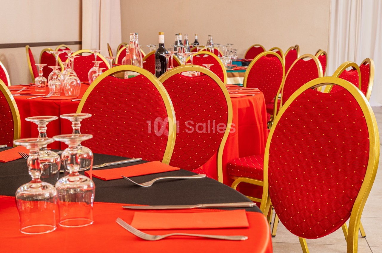 Salle de réception avec tables dressées, nappes rouges, chaises assorties et verres disposés pour un repas.