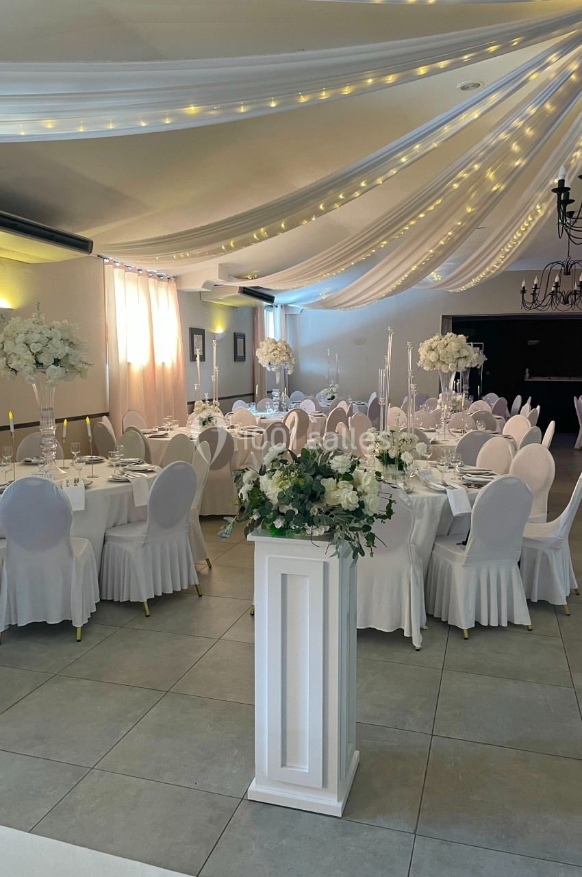 Salle de réception décorée avec des tables rondes, nappes blanches, compositions florales et guirlandes lumineuses au…