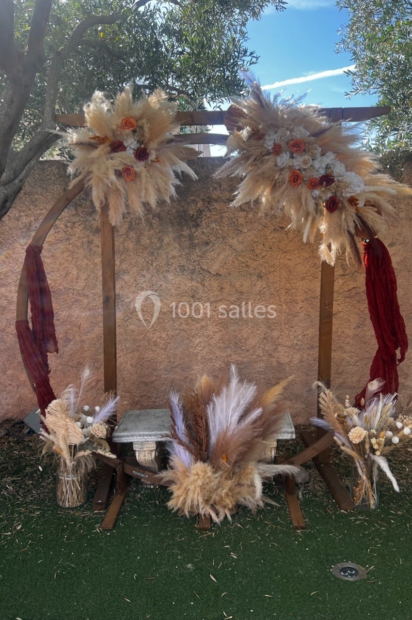 Location salle Cuers (Var) - L'atrium #13 Arche décorative en bois ornée de fleurs séchées et pampas, installée sur une pelouse devant un mur en pierre.