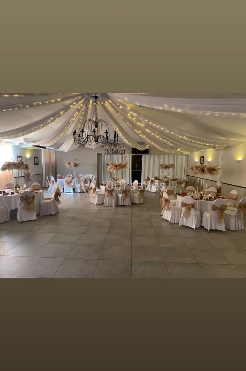 Location salle Cuers (Var) - L'atrium #5 Salle de réception décorée avec des tables rondes, nappes blanches, chaises ornées de nœuds beige et guirlandes lumineuses…