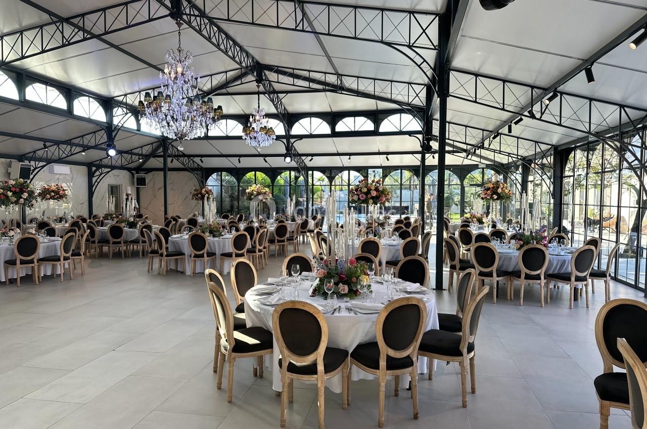 Salle de réception lumineuse avec tables rondes décorées de fleurs et chaises élégantes sous une verrière.