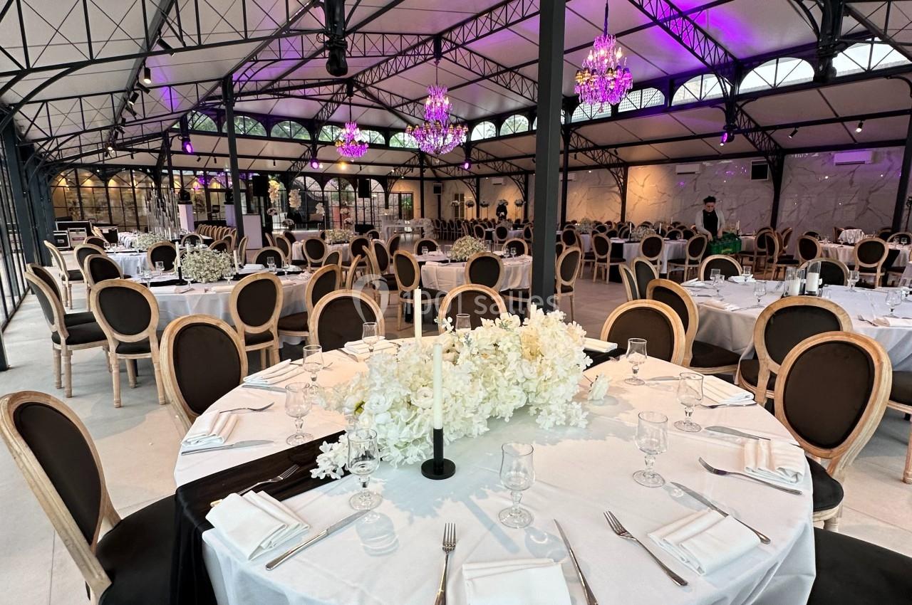 Salle de réception élégante avec tables rondes dressées, nappes blanches, chandeliers et éclairage violet.