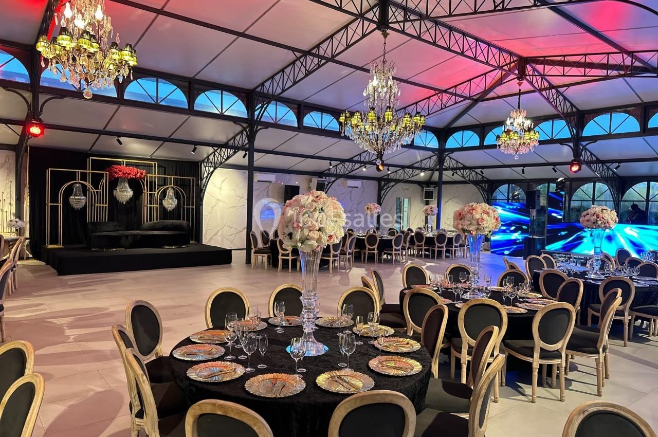Salle de réception élégante avec tables dressées, chandeliers suspendus et scène décorée en arrière-plan.