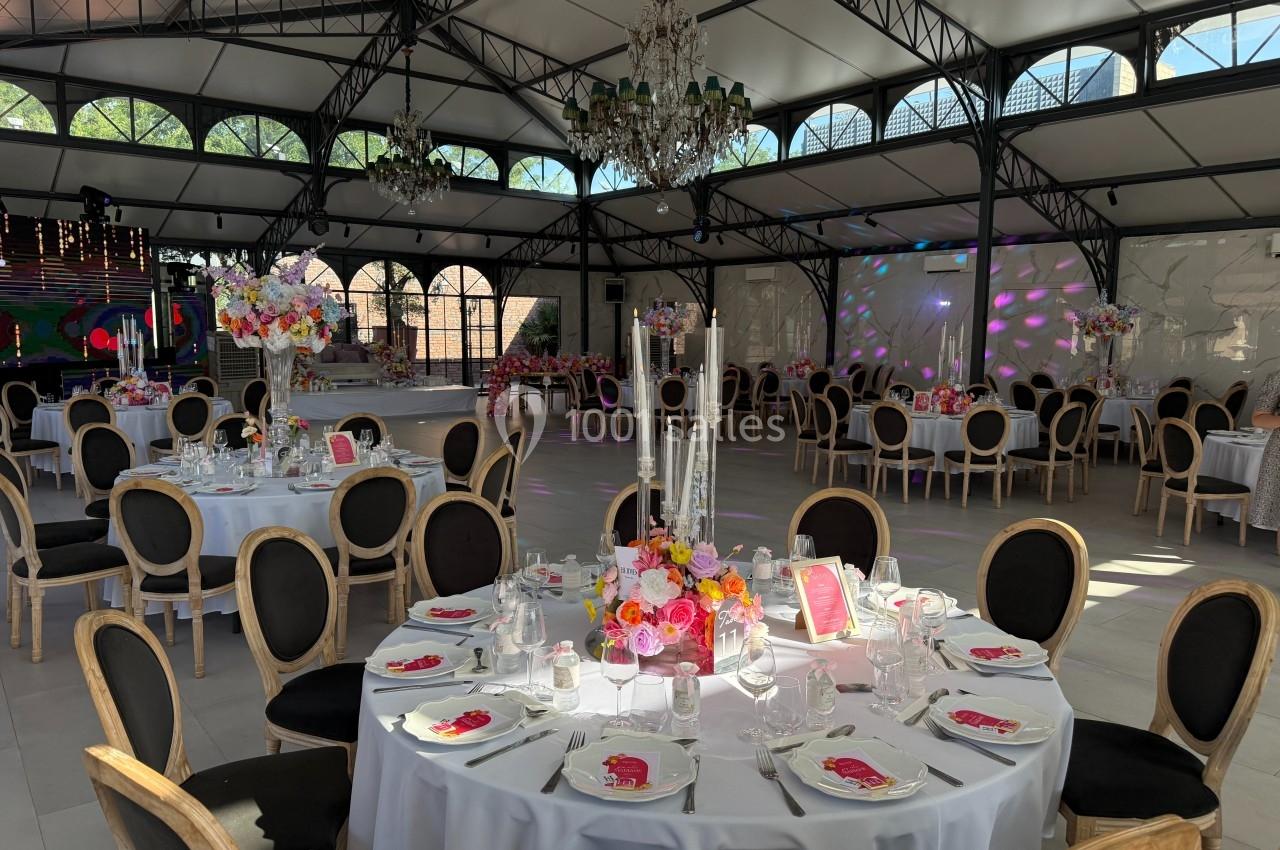 Salle de réception élégante avec tables rondes décorées de fleurs et chandeliers sous une verrière lumineuse.