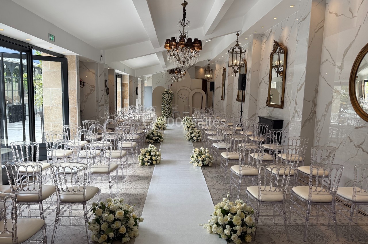 Salle de cérémonie décorée avec des chaises transparentes, un tapis blanc et des arrangements floraux blancs.