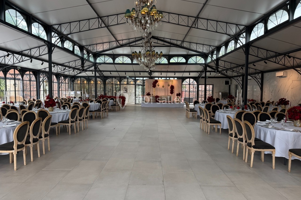 Salle de réception spacieuse avec tables dressées, chaises élégantes et scène décorée au fond sous un plafond vitré.