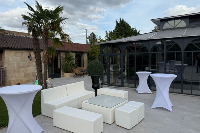 Terrasse extérieure avec tables, chaises, parasol, plantes et vue sur un bâtiment vitré sous un ciel dégagé.