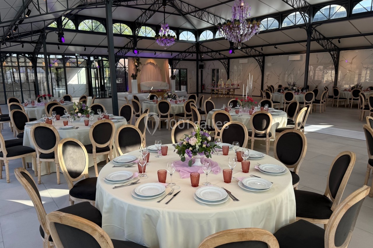 Salle de réception élégante avec tables rondes dressées, chaises noires et lustres suspendus sous une verrière.