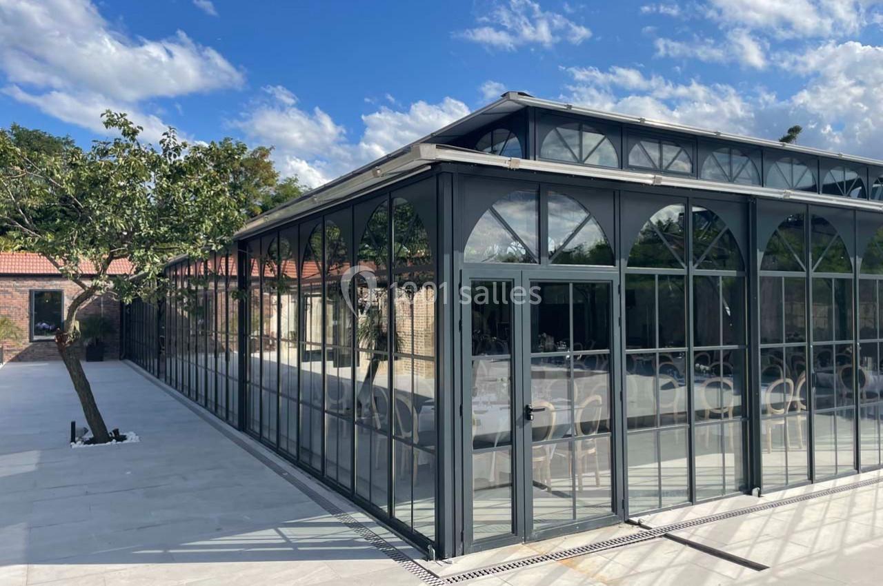 Verrière moderne en métal et verre avec des tables dressées à l'intérieur, entourée d'une terrasse pavée et d'arbres.