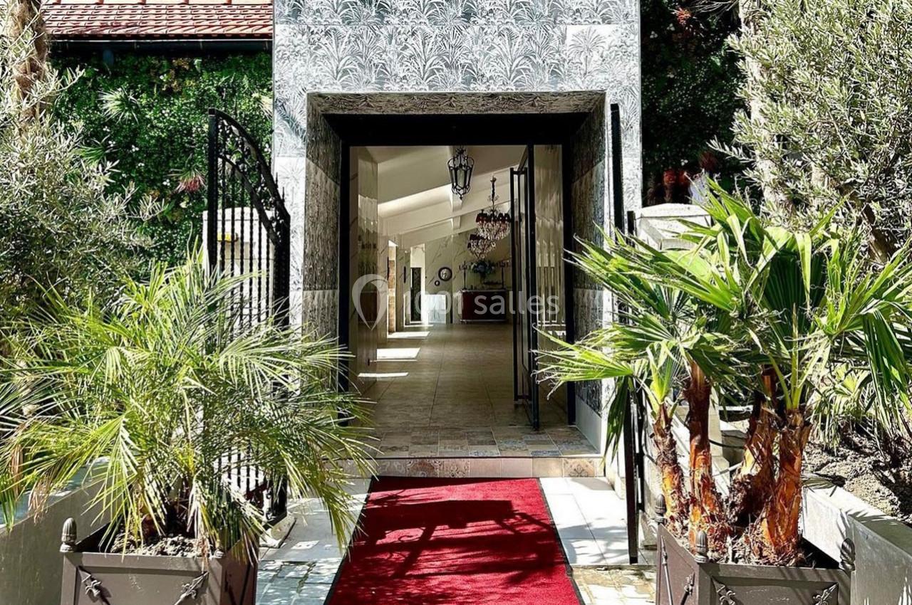 Entrée d'un bâtiment avec un tapis rouge, encadrée de plantes en pot et d'un portail en fer forgé.