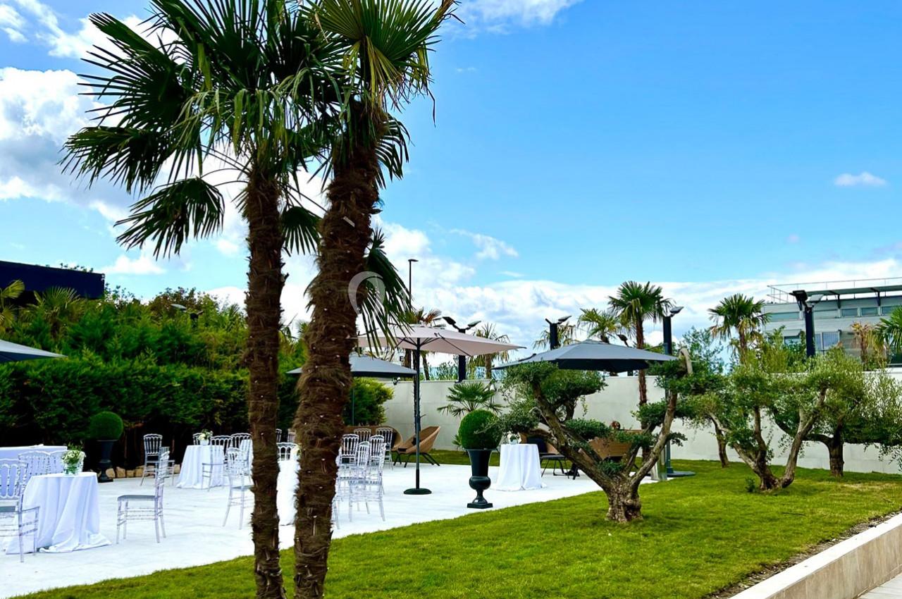 Jardin avec pelouse, palmiers, tables dressées et parasols noirs sous un ciel partiellement nuageux.